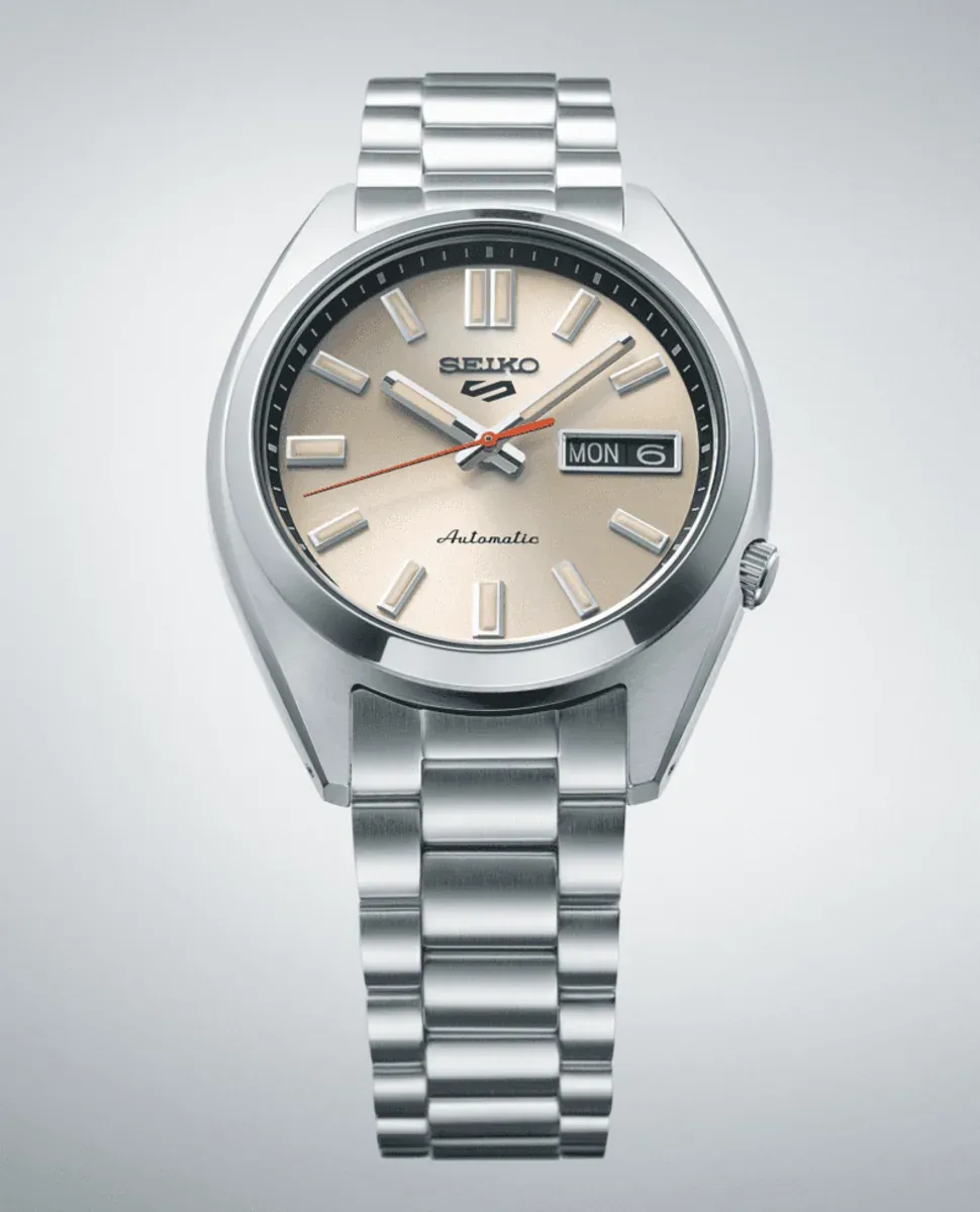 Vista frontal completa del reloj con esfera beige y brazalete de acero sobre fondo gris degradado.