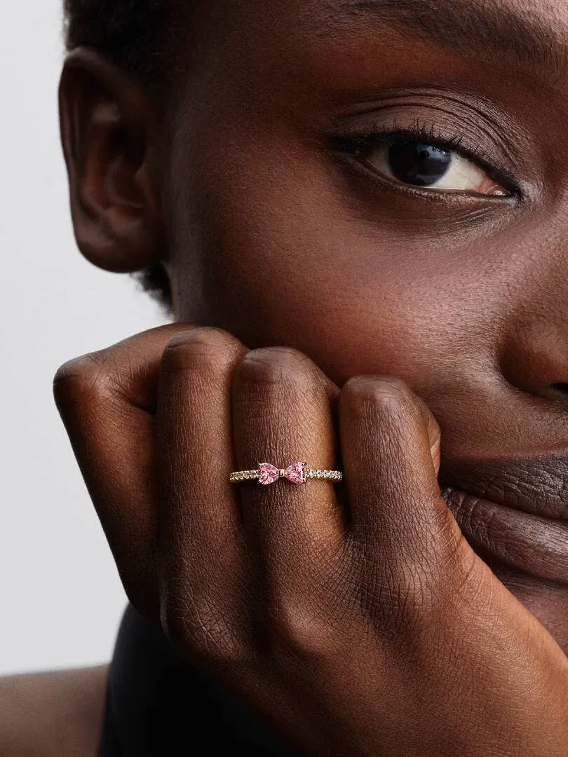 Primer plano del anillo de lazo rosa en el dedo, toma lateral con rostro de modelo desenfocado al fondo.