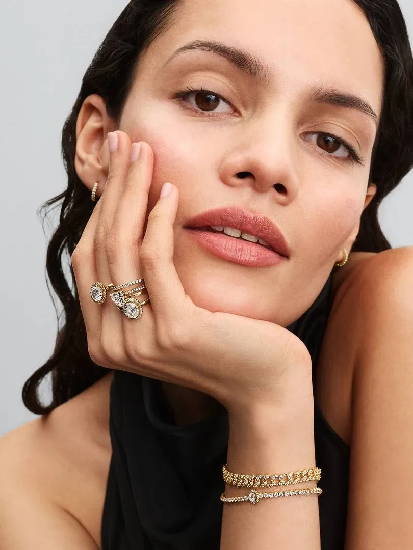 Primer plano de mano con anillos y brazaletes dorados, modelo apoyada en la cara sobre fondo neutro.