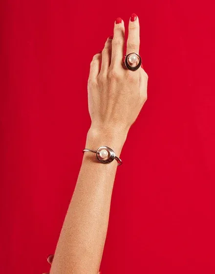Bras et main portant un bracelet en acier et une bague en perle, sur fond rouge vif, vue verticale.