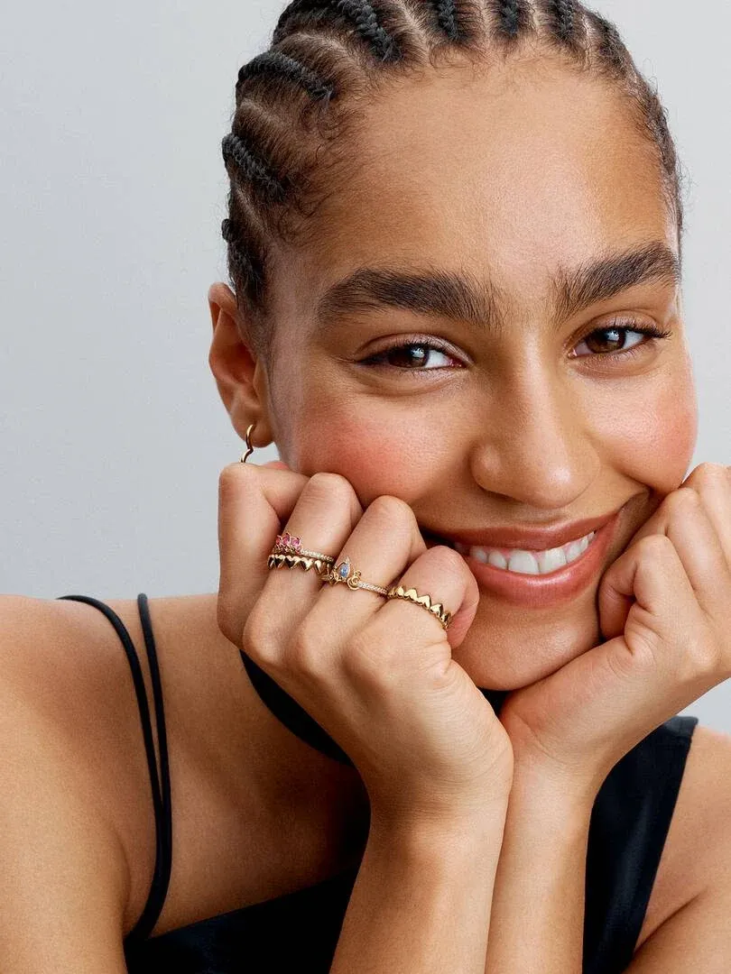 Primer plano de manos en la cara mostrando el anillo tipo tiara con gemas rosas, fondo neutro claro.