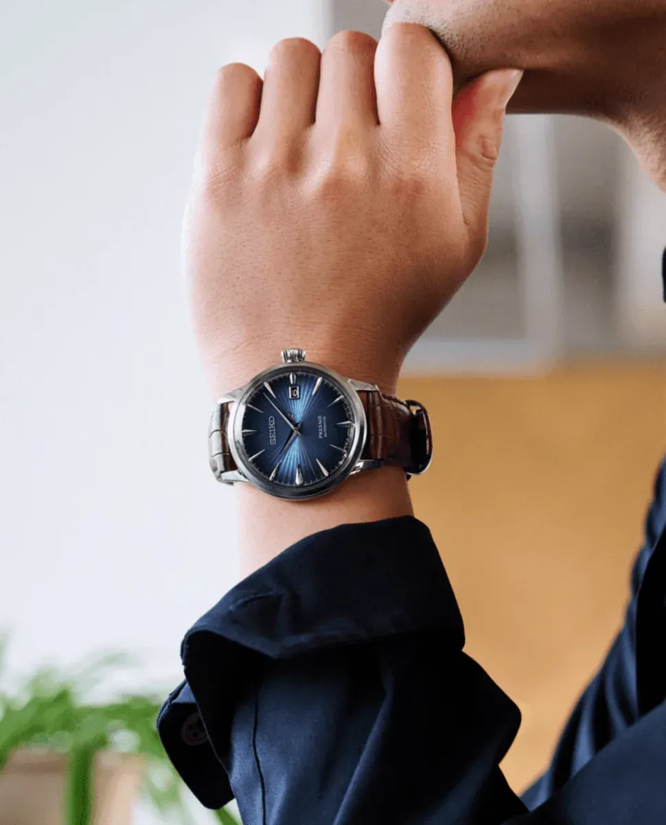 Reloj puesto en muñeca masculina, plano medio corto, fondo interior desenfocado y camisa azul oscuro.