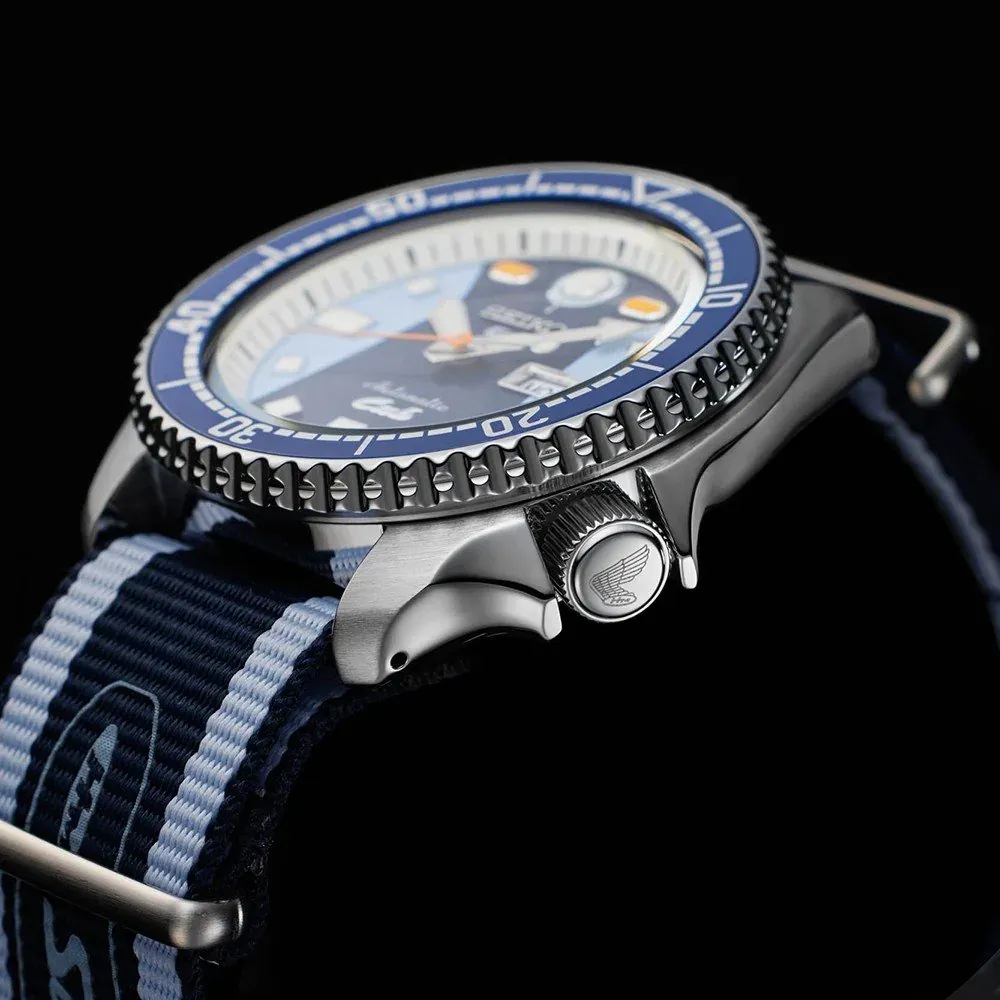 Vista lateral en ángulo del reloj sobre fondo negro, detalle de la corona grabada y la correa de tela a rayas