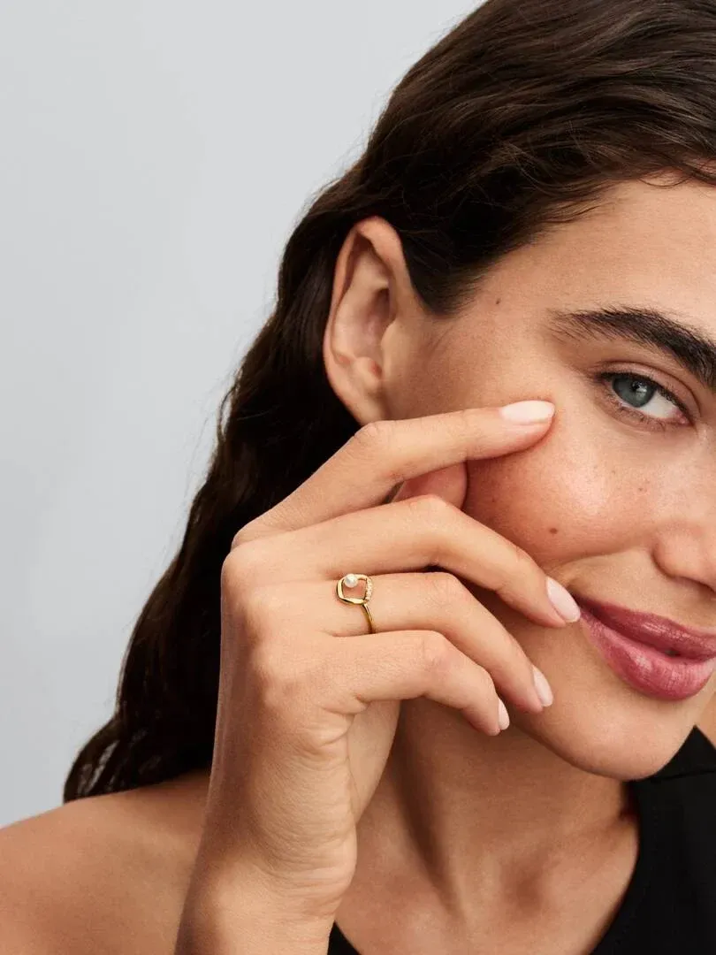 Primer plano de anillo con perla en dedo medio, mano apoyada en la mejilla, fondo neutro claro.