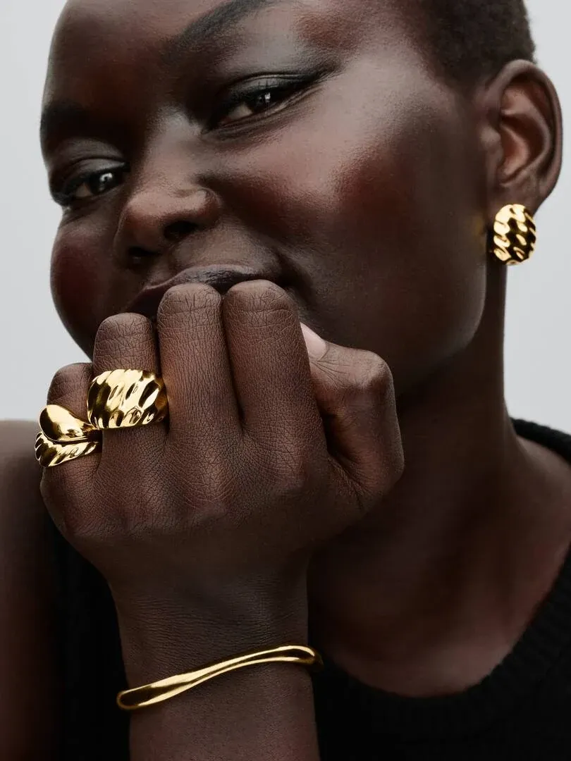 Primer plano de mano con anillo ondulado dorado y pendiente a juego, sobre modelo, fondo neutro.