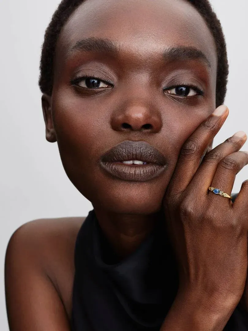 Primer plano de mano femenina apoyada en la cara, luciendo el anillo con gema azul sobre fondo neutro.