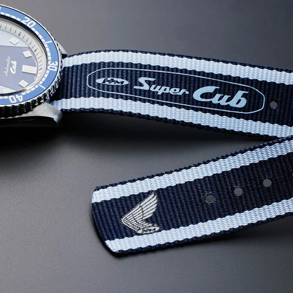 Primer plano de la correa de tela a rayas azules y blancas con inscripción 'Super Cub', fondo gris neutro