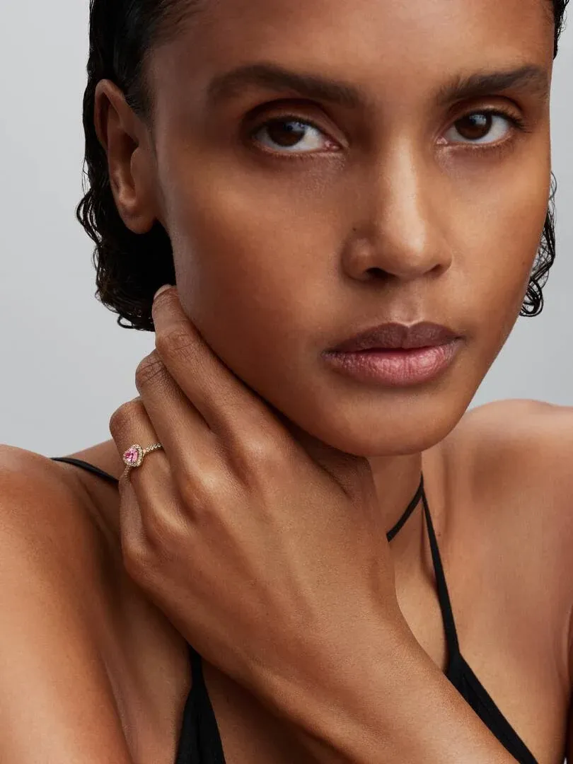 Primer plano de anillo con corazón rosa en dedo anular, modelo posando con la mano cerca del rostro.