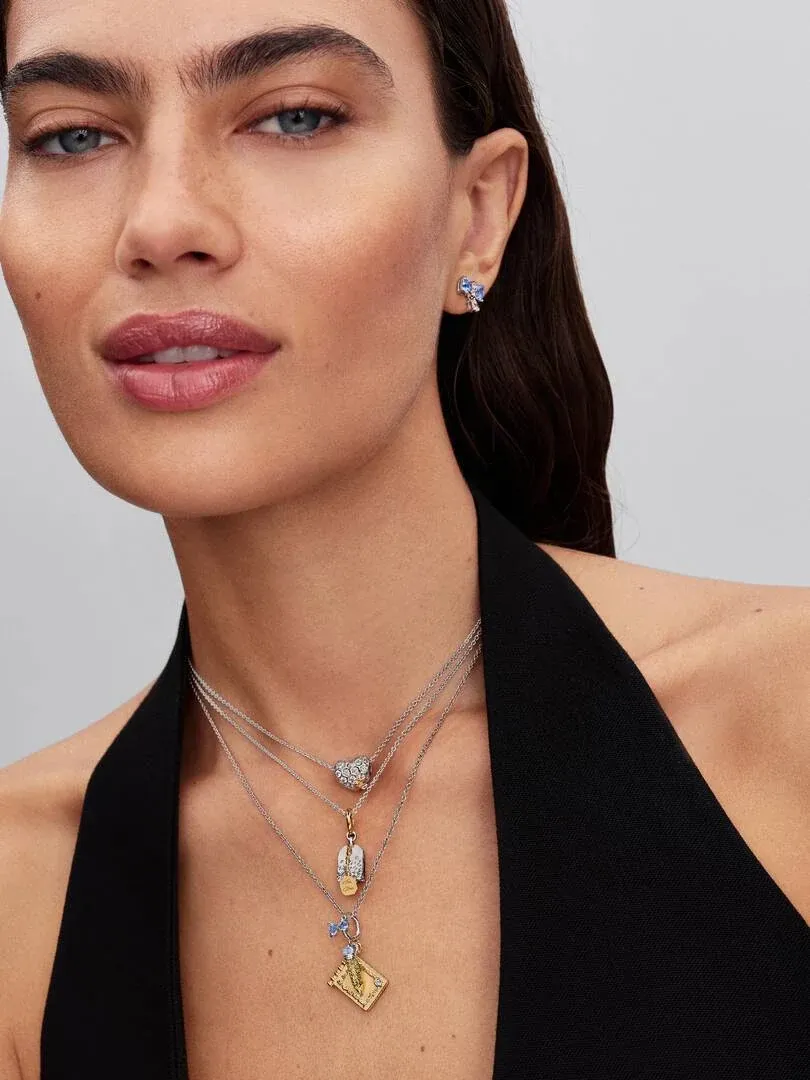 Nahaufnahme eines Models mit mehrlagigen Halsketten und blauen Ohrsteckern, Frontansicht vor neutralem Hintergrund.