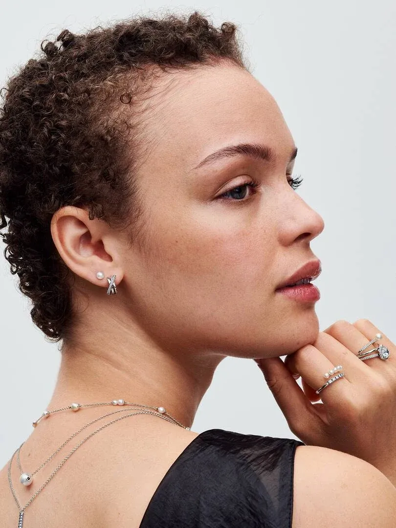 Primer plano de perfil de modelo luciendo pendientes de aro cruzados y collar de perlas sobre fondo neutro.