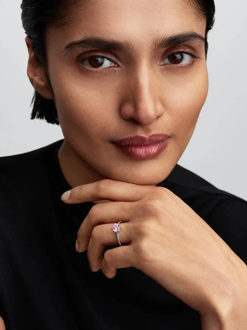 Primer plano de anillo con piedra fucsia en dedo anular, mano apoyada en la barbilla, fondo neutro.