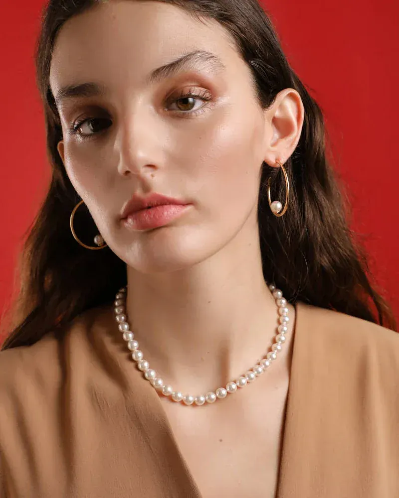 Plano medio de modelo luciendo collar de perlas y pendientes aros, fondo rojo sólido.