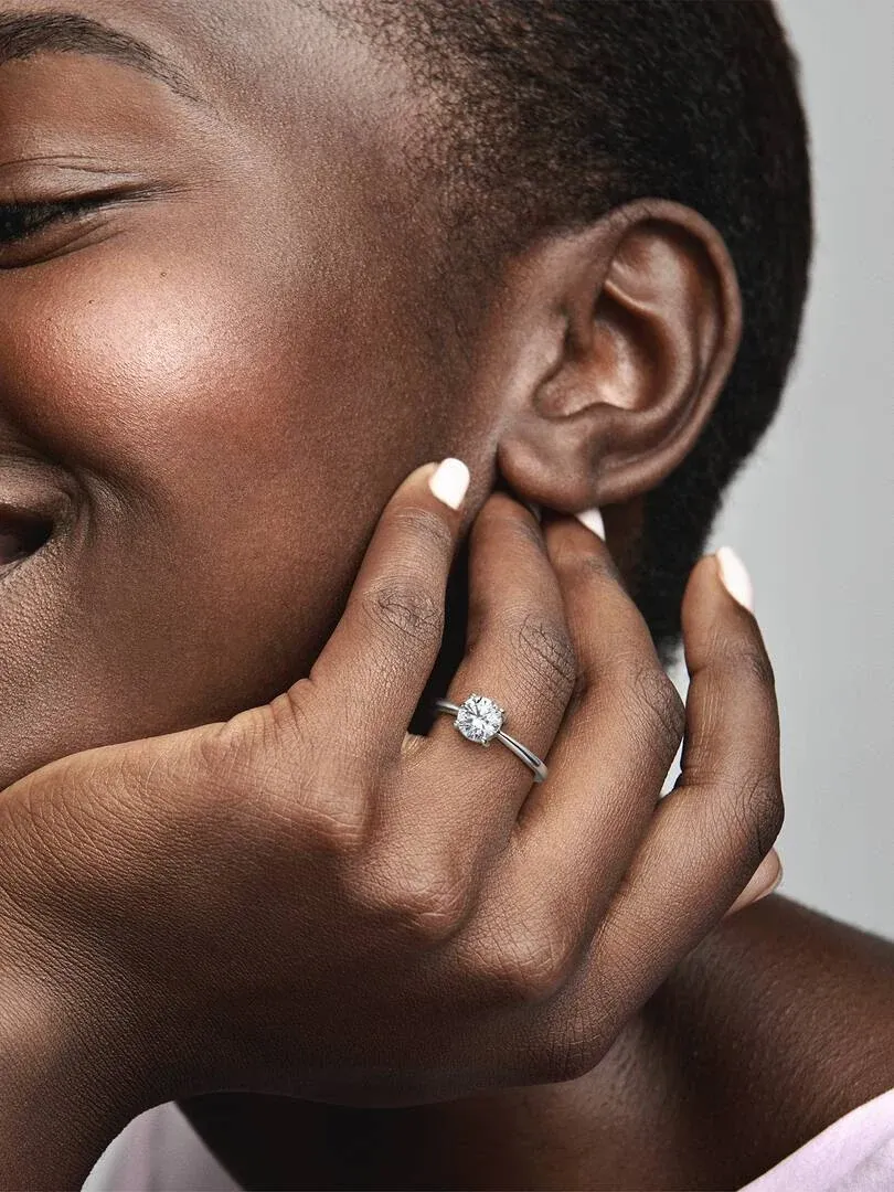 Close-up of the solitaire ring on the ring finger, hand resting near the face, neutral background.