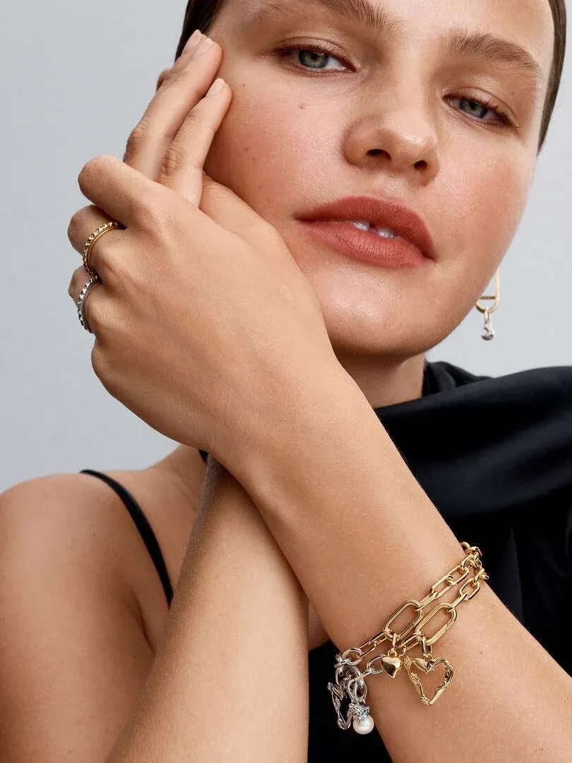 Nahaufnahme eines Models mit einem goldenen Gliederarmband mit Charms, die die Hand ans Gesicht hält.