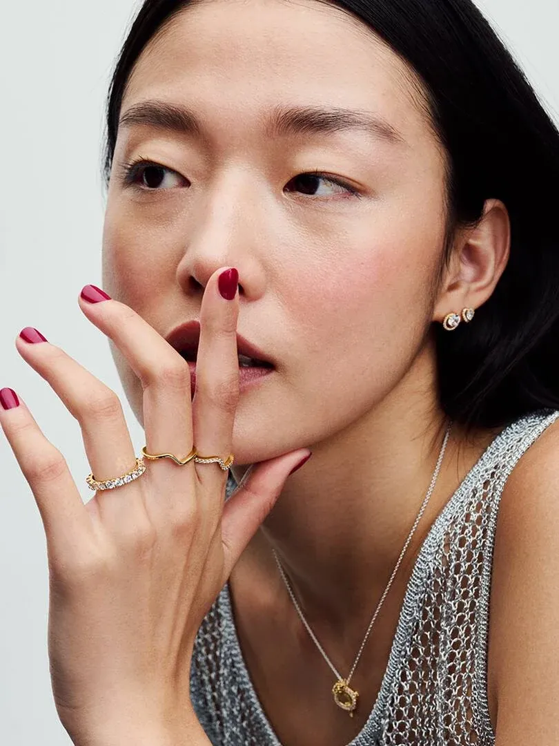 Primer plano de mano con anillos dorados ondulados y pendientes, modelo con fondo neutro.