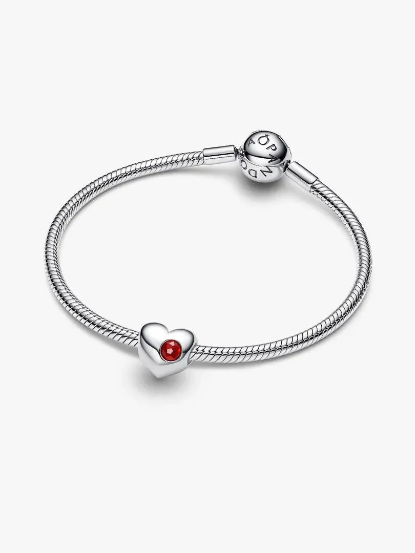Charm de corazón con gema roja sobre brazalete de plata, vista frontal en ángulo, fondo blanco.