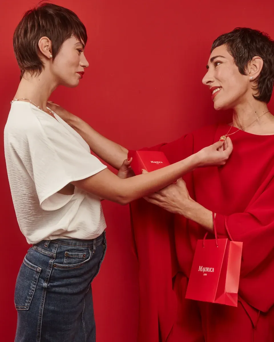 Deux femmes sur fond rouge, l'une passant le collier à l'autre ; sac cadeau Majorica visible.