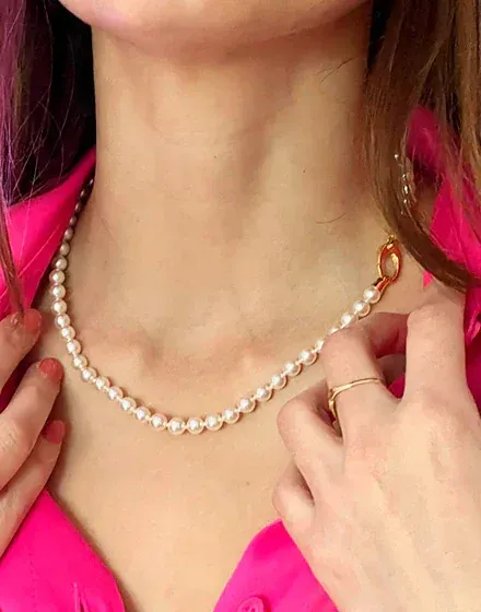 Primer plano del collar de perlas puesto en el cuello, con camisa rosa vibrante y detalle de anillo dorado.