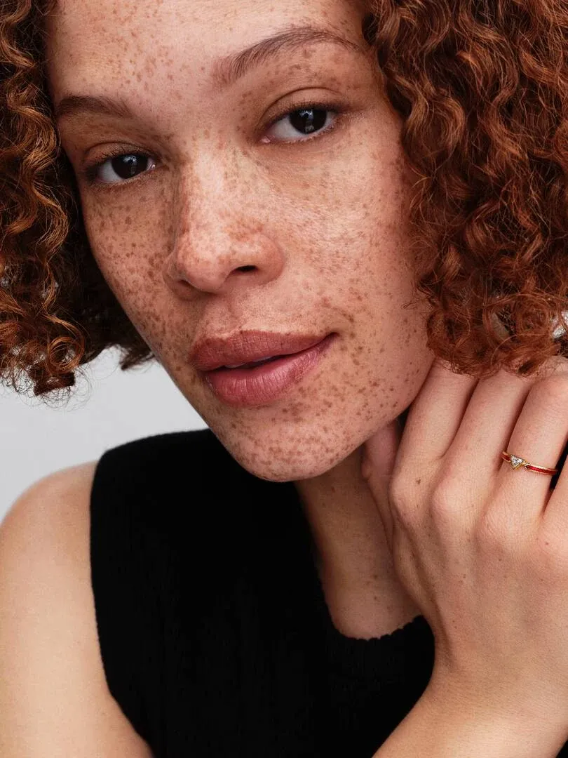 Primer plano de anillo dorado con gema roja en el dedo anular, mano apoyada en la mejilla.