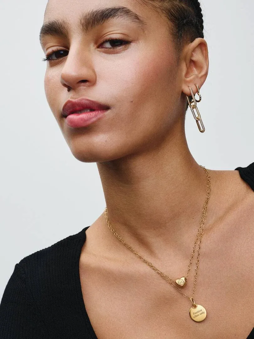 Gros plan d'un modèle portant un collier à double chaîne avec pendentif rond gravé et boucles d'oreilles créoles.