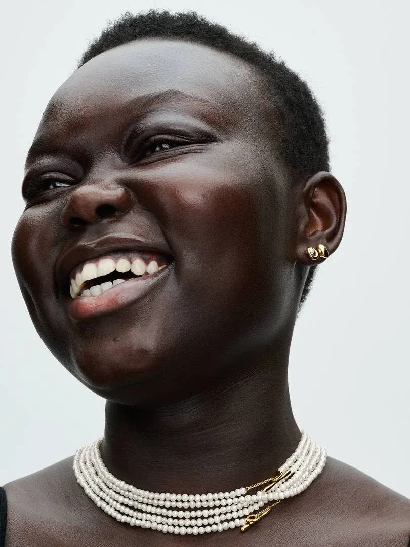 Primer plano de modelo sonriendo con collar de perlas blancas y pendientes dorados, fondo neutro claro.