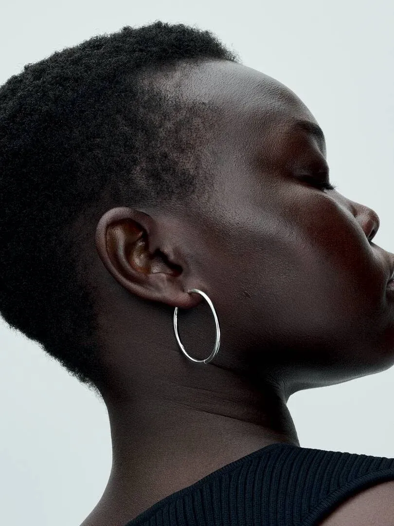 Gros plan de profil d'une boucle d'oreille créole en argent portée à l'oreille, fond neutre clair.