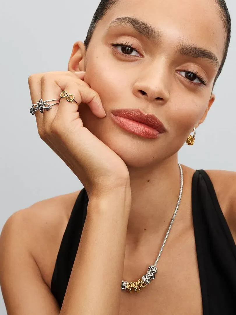 Primer plano de modelo con collar, pendientes y anillos de nudo infinito sobre fondo gris neutro.