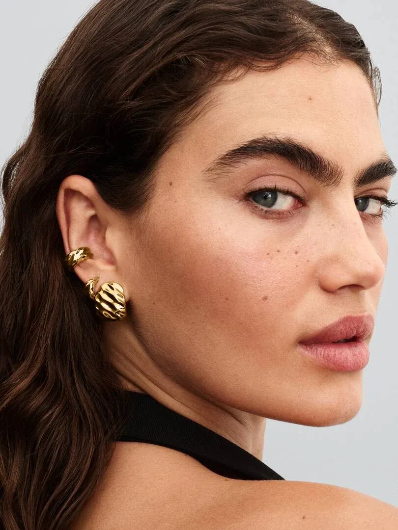 Gros plan latéral de boucles d'oreilles créoles dorées ondulées portées à l'oreille, fond neutre.