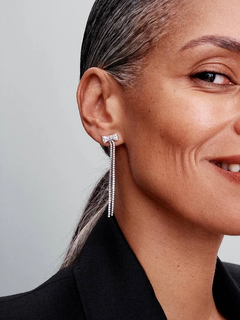 Gros plan latéral de boucle d'oreille pendante avec nœud et pavé de zircons portée à l'oreille, fond neutre.