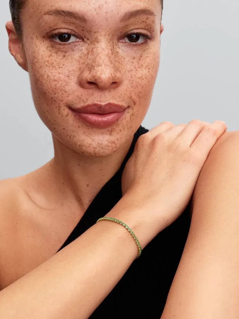 Primer plano de pulsera tenis verde en la muñeca, modelo con pecas y fondo neutro.