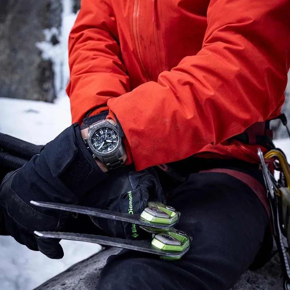 Reloj puesto en muñeca con guantes negros y chaqueta roja, sosteniendo piolets en un entorno de montaña nevada.