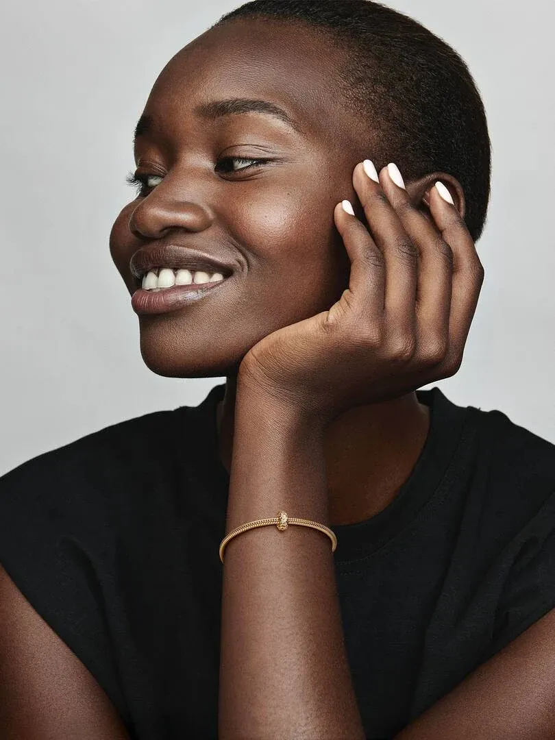 Gros plan d'un bracelet doré au poignet, modèle souriant de profil sur fond gris neutre.