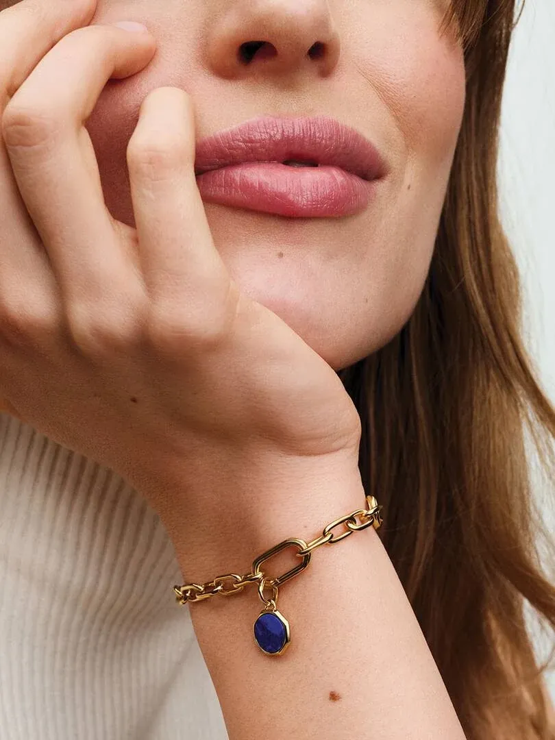 Nahaufnahme eines Handgelenks mit einem goldenen Gliederarmband und blauem Charm, an das Gesicht eines Models gelehnt.