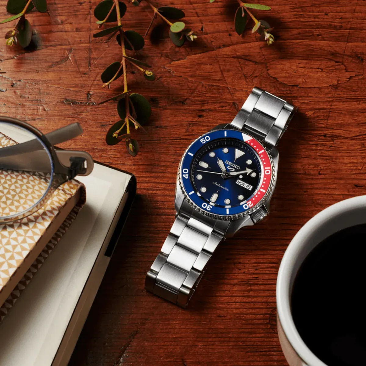 Reloj sobre mesa de madera en composición cenital con cuaderno, gafas, taza de café y hojas decorativas.