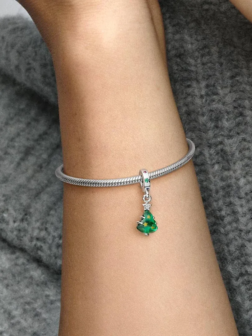 Close-up of green Christmas tree hanging charm on a silver bracelet, worn on wrist against grey background.