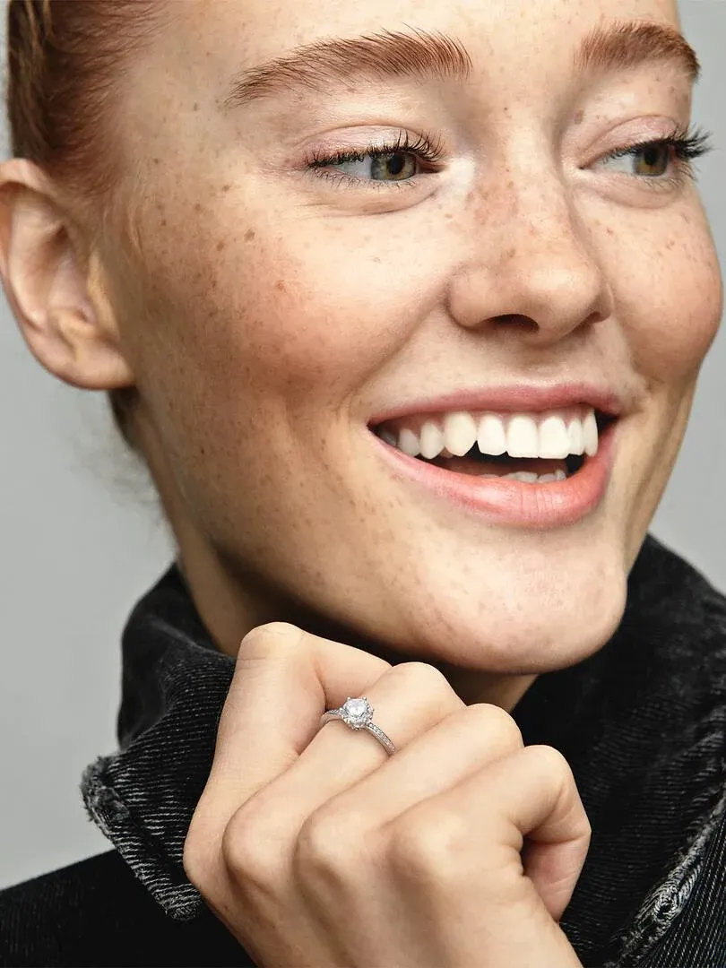 Primer plano de anillo con circonita sobre dedo, modelo sonriente con fondo desenfocado.