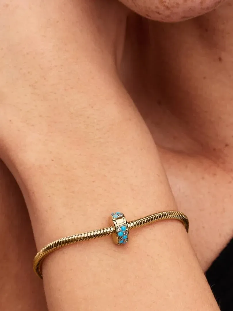 Nahaufnahme des blauen Clip-Charms an einem goldenen Armband, getragen am Handgelenk.