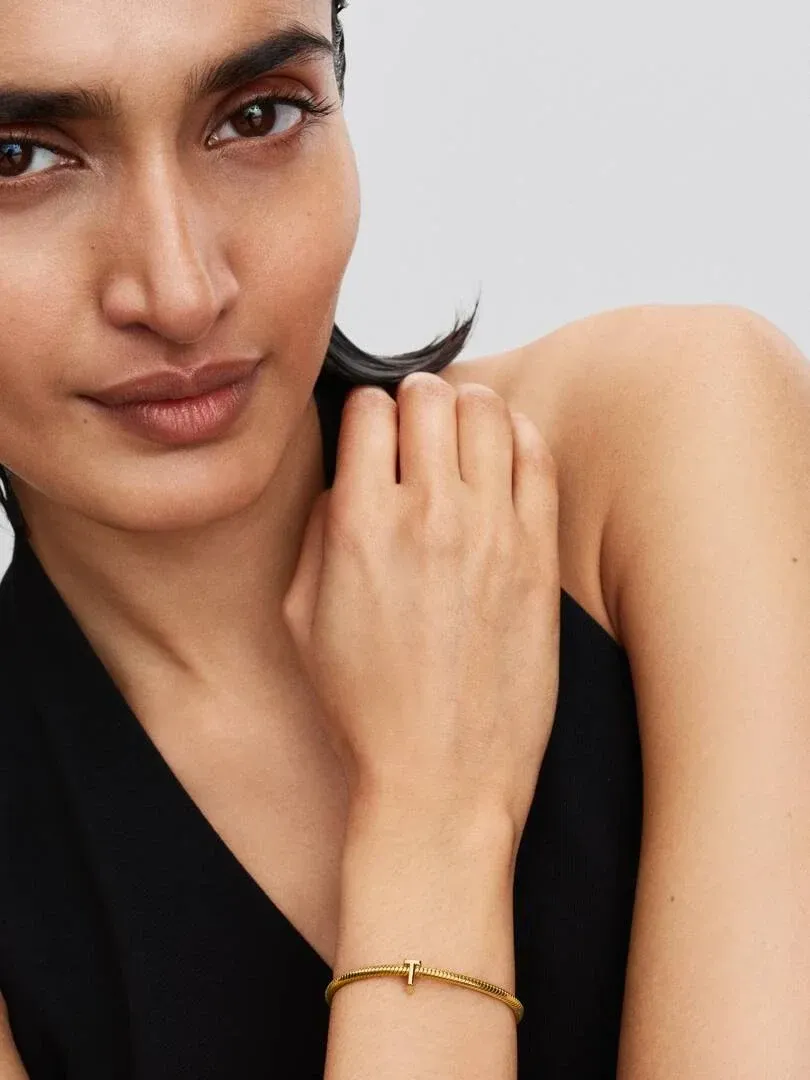 Gros plan du poignet portant le charm lettre T doré, modèle en haut noir sur fond neutre.