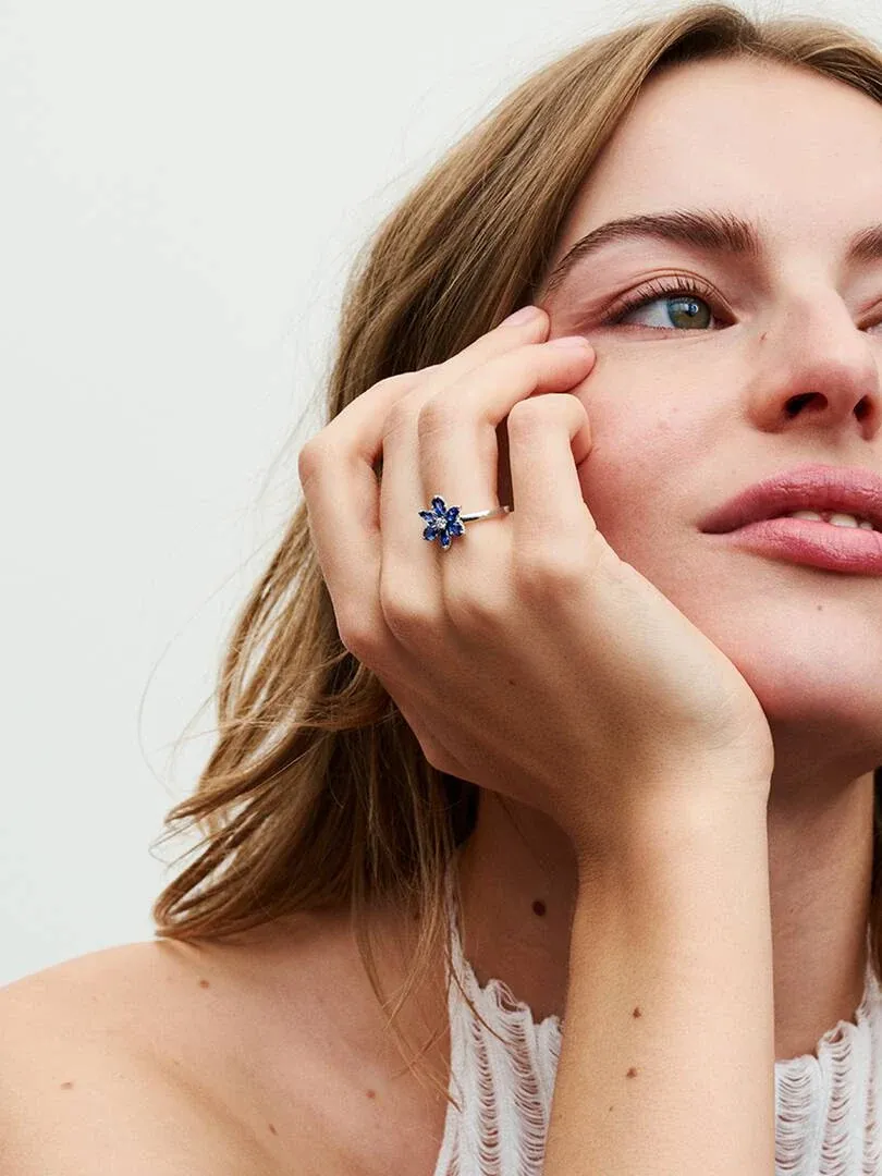 Close-up of blue floral ring on finger, model resting face on hand, white background.