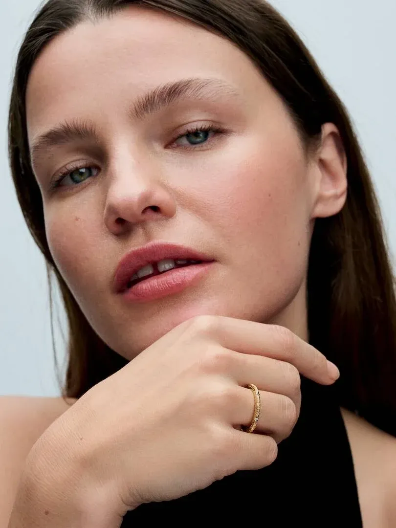 Primer plano de modelo con el anillo dorado en el dedo, toma lateral cerca del rostro sobre fondo claro.