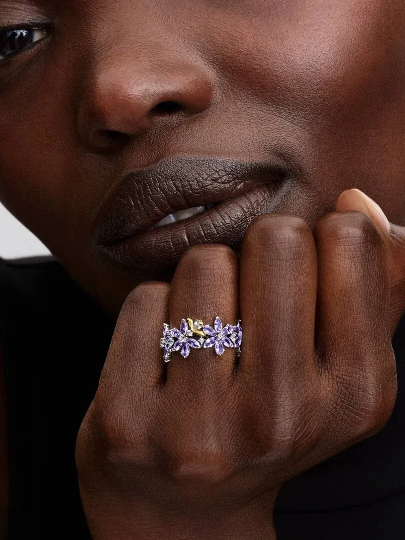 Primer plano del anillo con flores moradas y abeja en el dedo de una modelo, toma lateral sobre fondo oscuro.