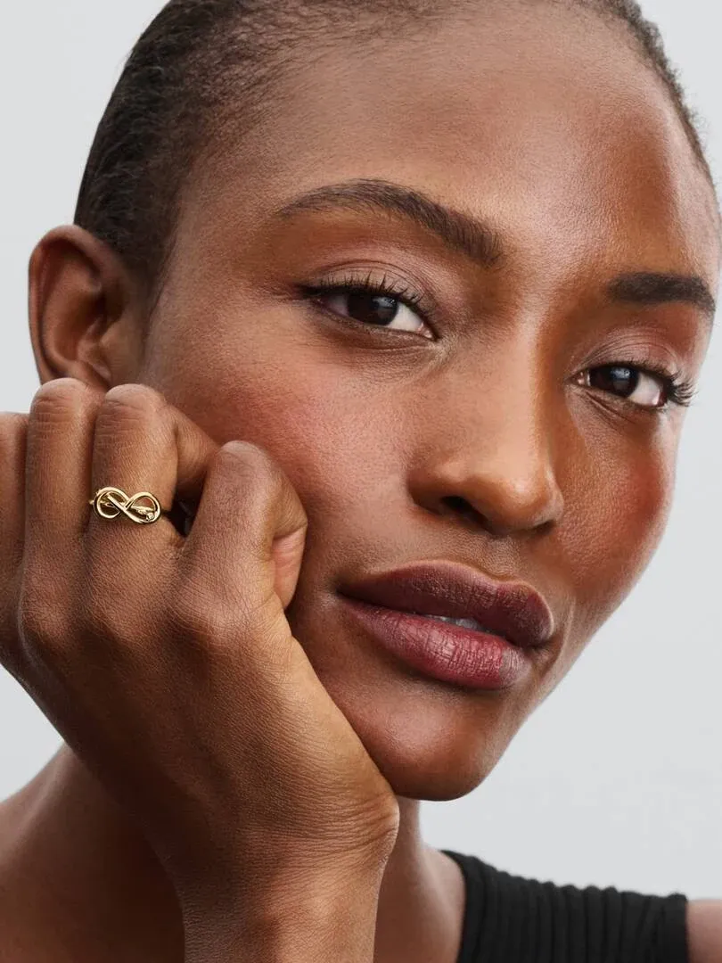 Primer plano del anillo de nudo infinito en el dedo, modelo apoyando la cara sobre su mano, fondo neutro.