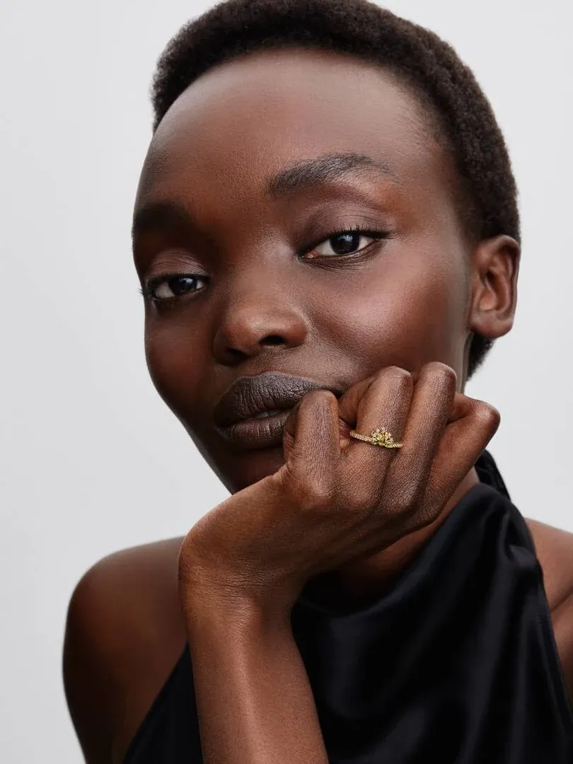 Primer plano de la mano apoyada en el mentón, mostrando el anillo dorado con gemas verdes sobre fondo neutro.