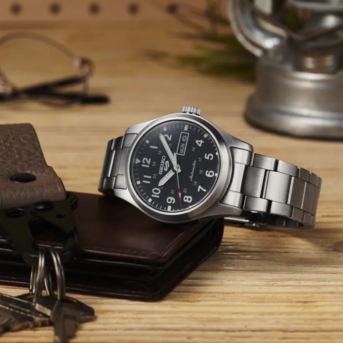 Reloj apoyado sobre una superficie de madera junto a una cartera de cuero y llaves, fondo desenfocado.