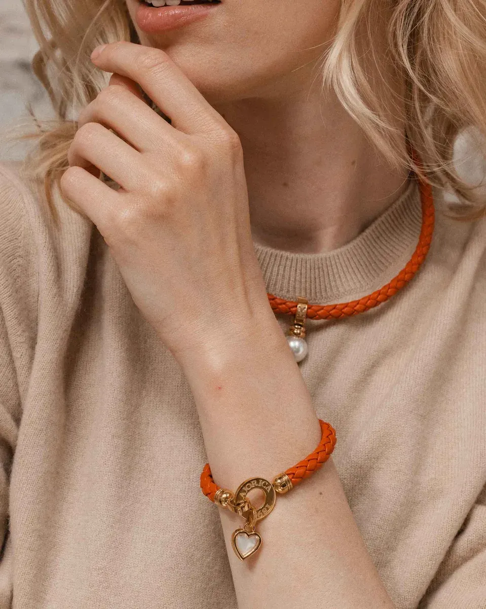 Primer plano de colgante de corazón y pulsera trenzada naranja sobre piel, toma lateral con fondo desenfocado.