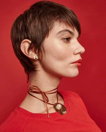 Perfil de modelo con pendientes dorados y collar de perla sobre fondo rojo vibrante.