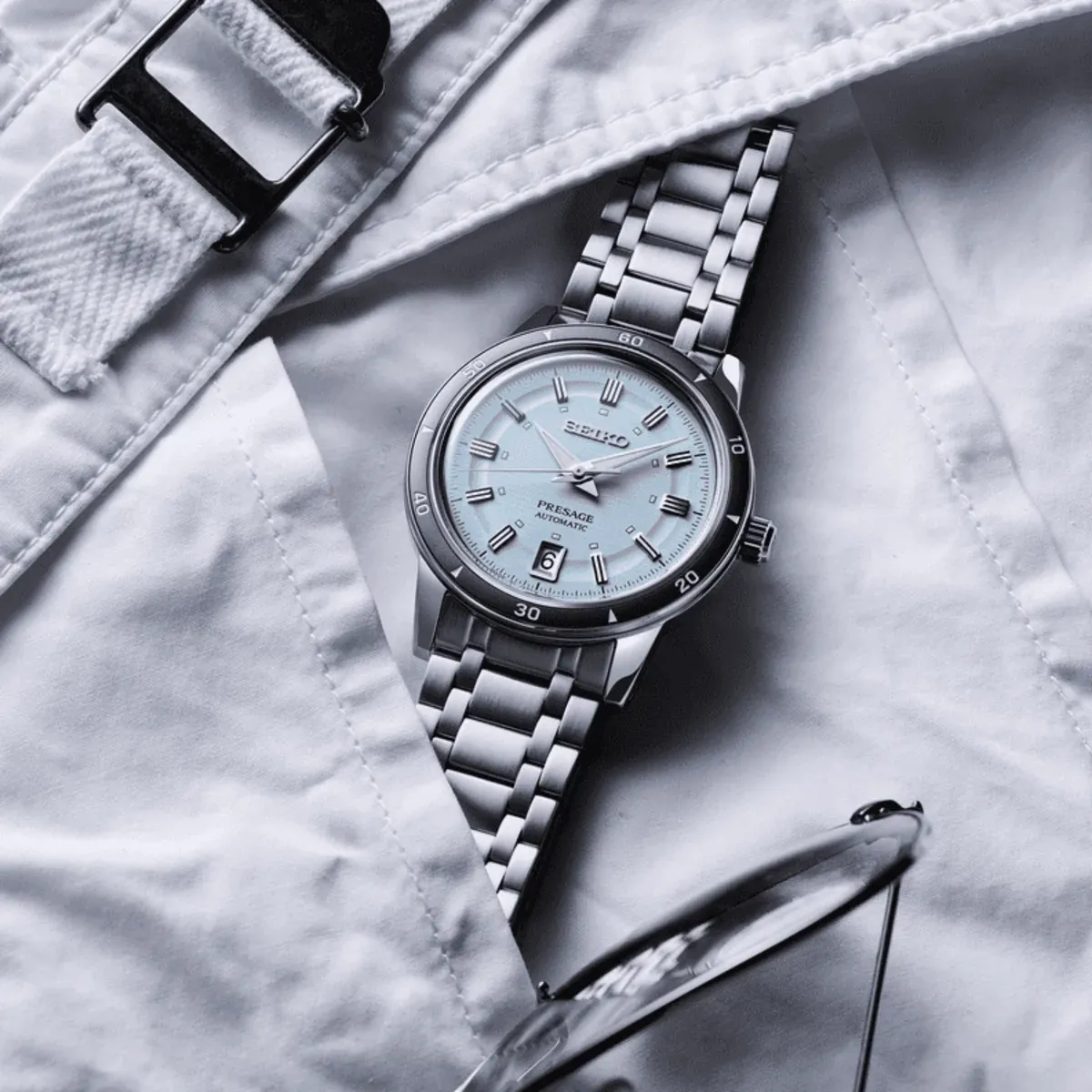 Reloj apoyado sobre tela blanca junto a un cinturón, vista cenital en tonos fríos y desaturados.