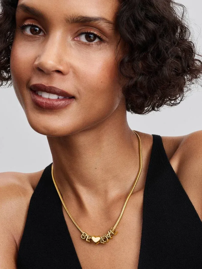 Close-up van gouden letterketting om de hals van een model, neutrale wazige achtergrond.