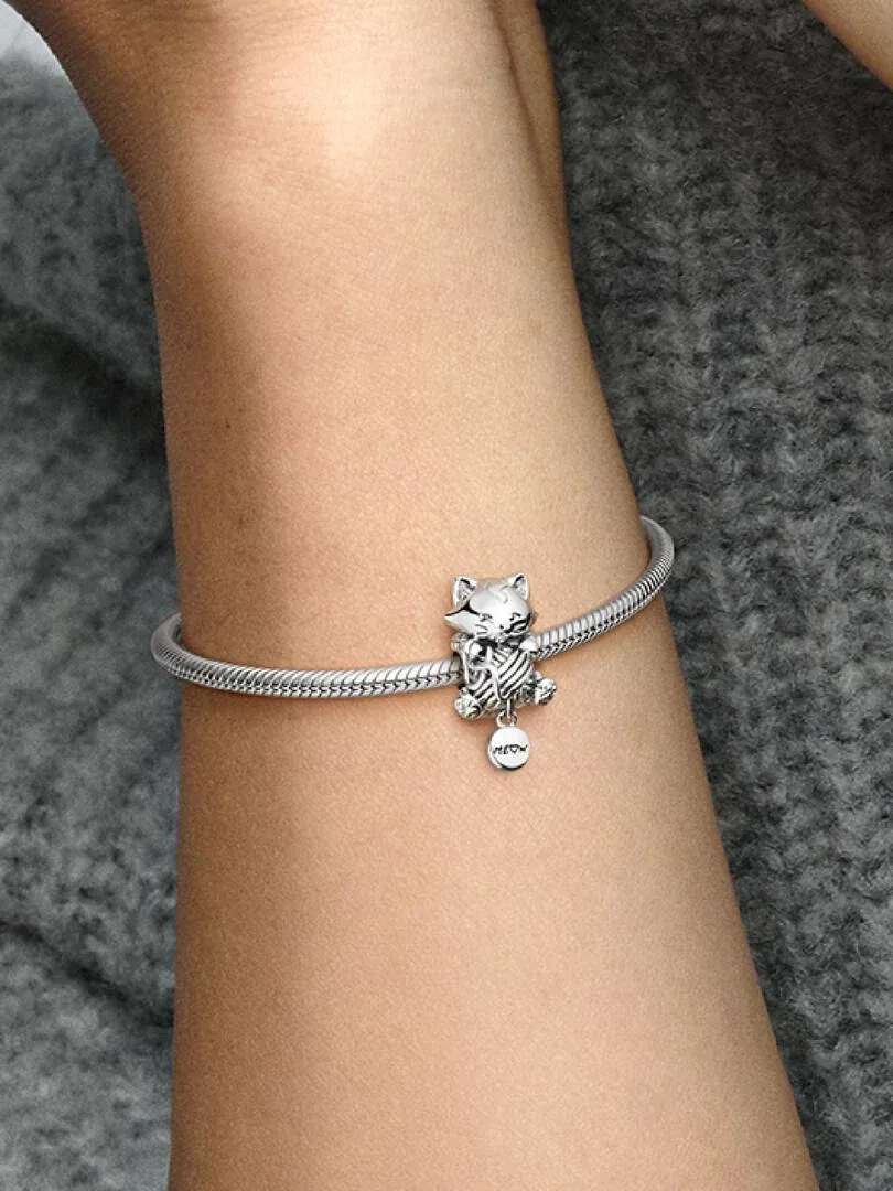 Primer plano del charm de gatito en una pulsera de plata sobre la muñeca, toma lateral con fondo gris desenfocado.