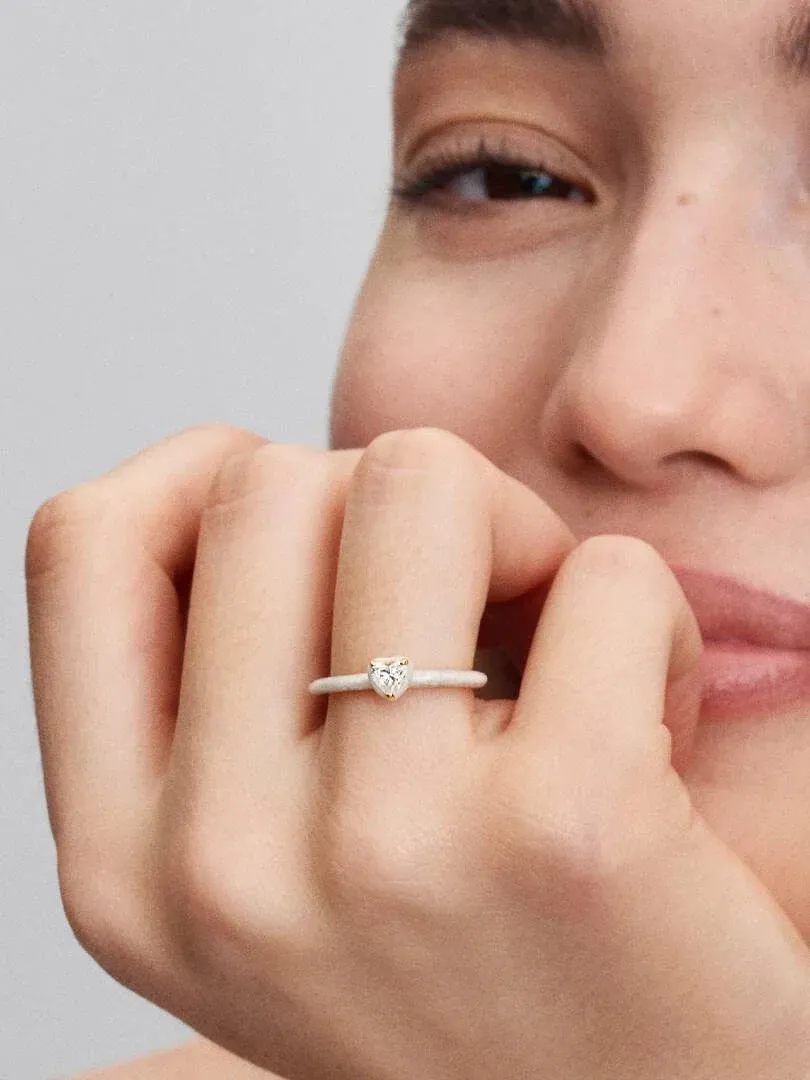 Primer plano del anillo con gema en forma de corazón puesto en el dedo, rostro de modelo desenfocado al fondo.
